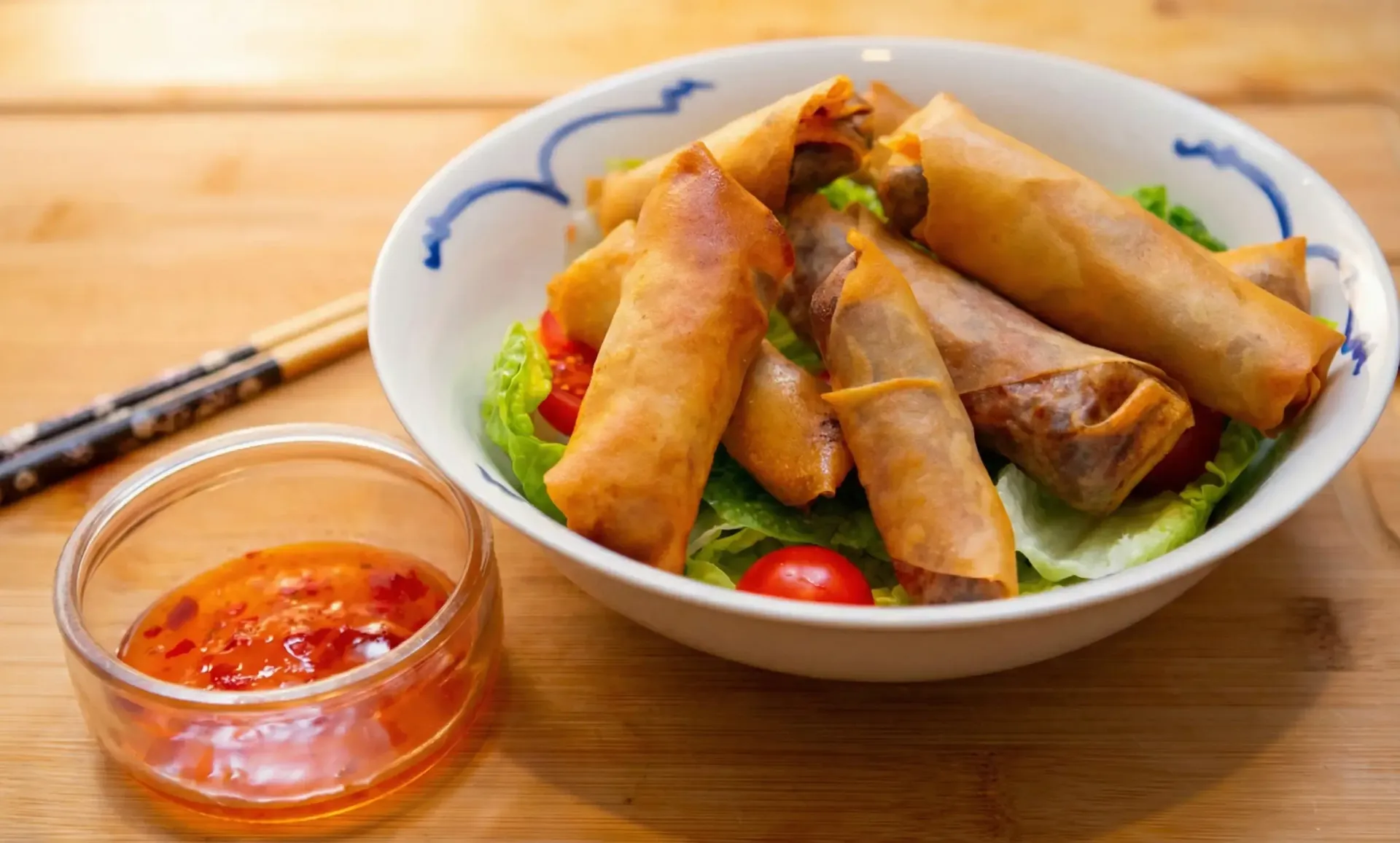Spring rolls in a bowl with dipping sauce and chopsticks on a wooden surface.