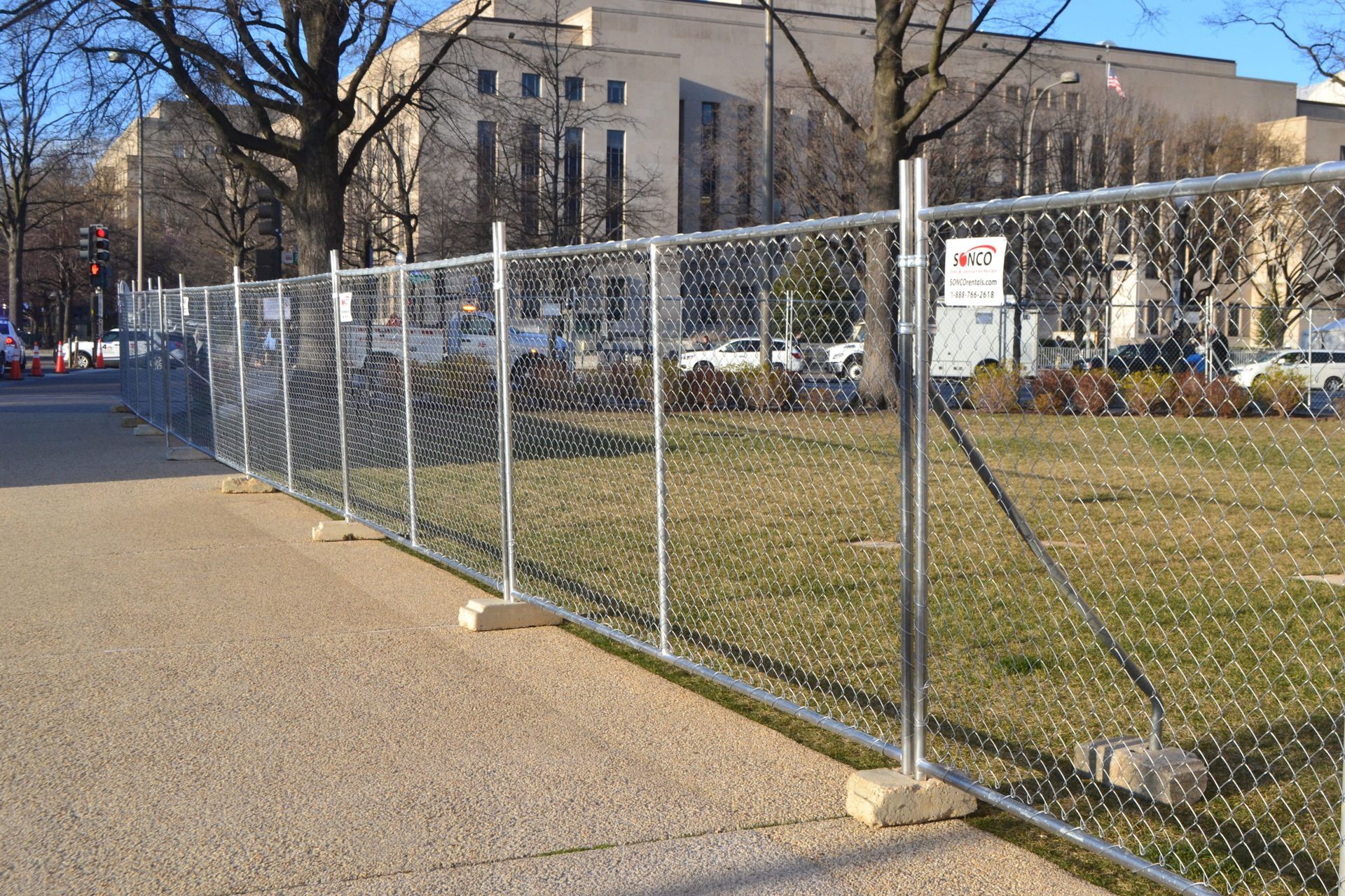 Chain-link fence along a sidewalk beside a grassy field and brick building on a sunny day