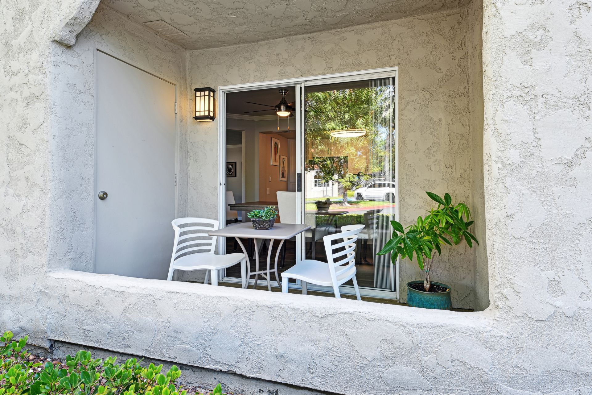 Patio with table, chairs, and potted plant, beside a sliding glass door and a solid wall.