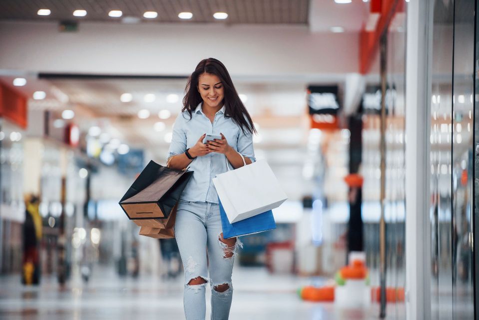 A woman is walking through a mall holding shopping bags and looking at her phone.