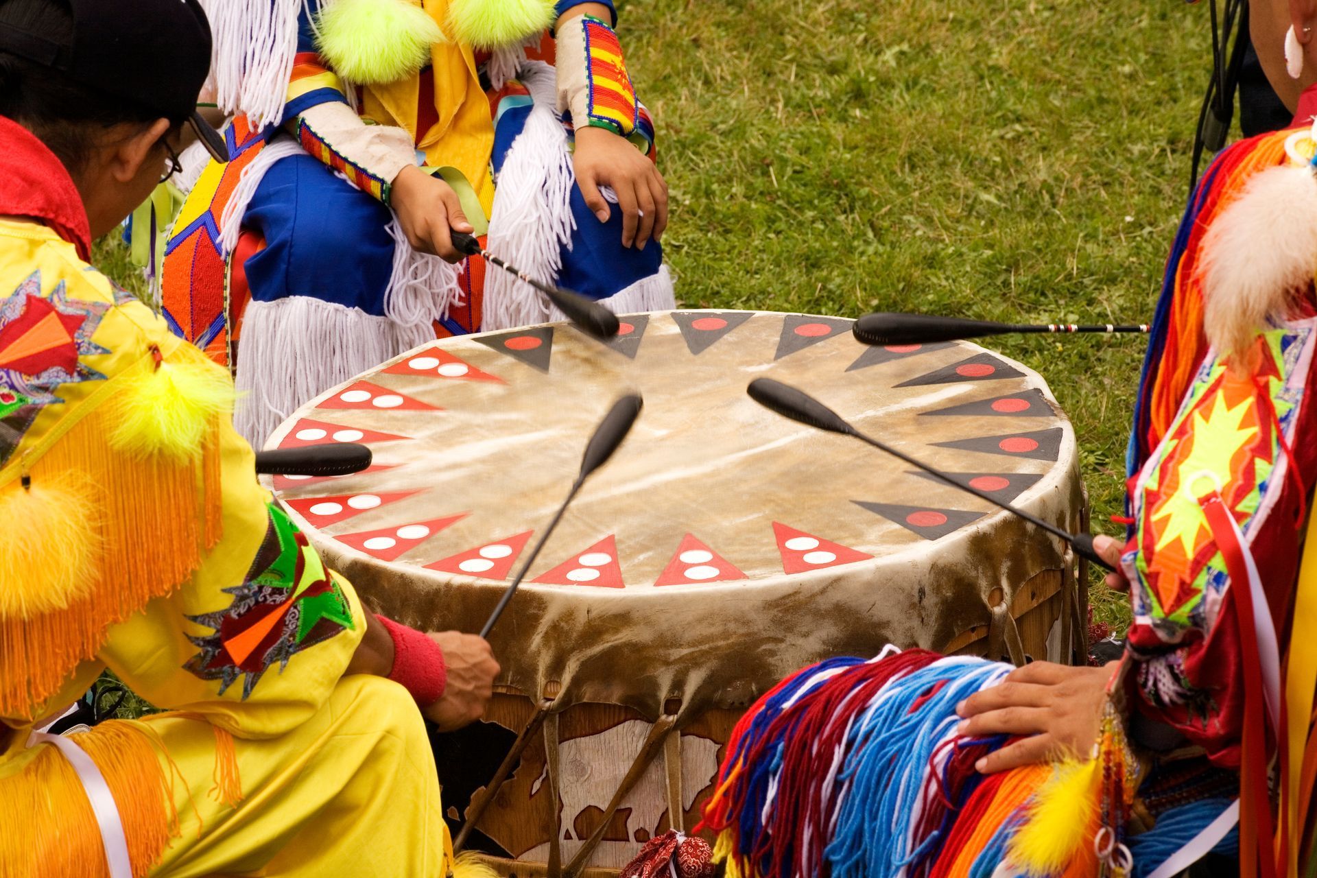 People in colorful Native American regalia playing a large drum with several drumsticks.