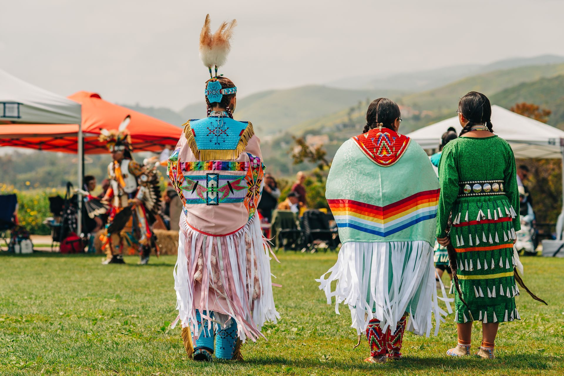 Three Native American dancers in colorful regalia at an outdoor event; mountains in background.