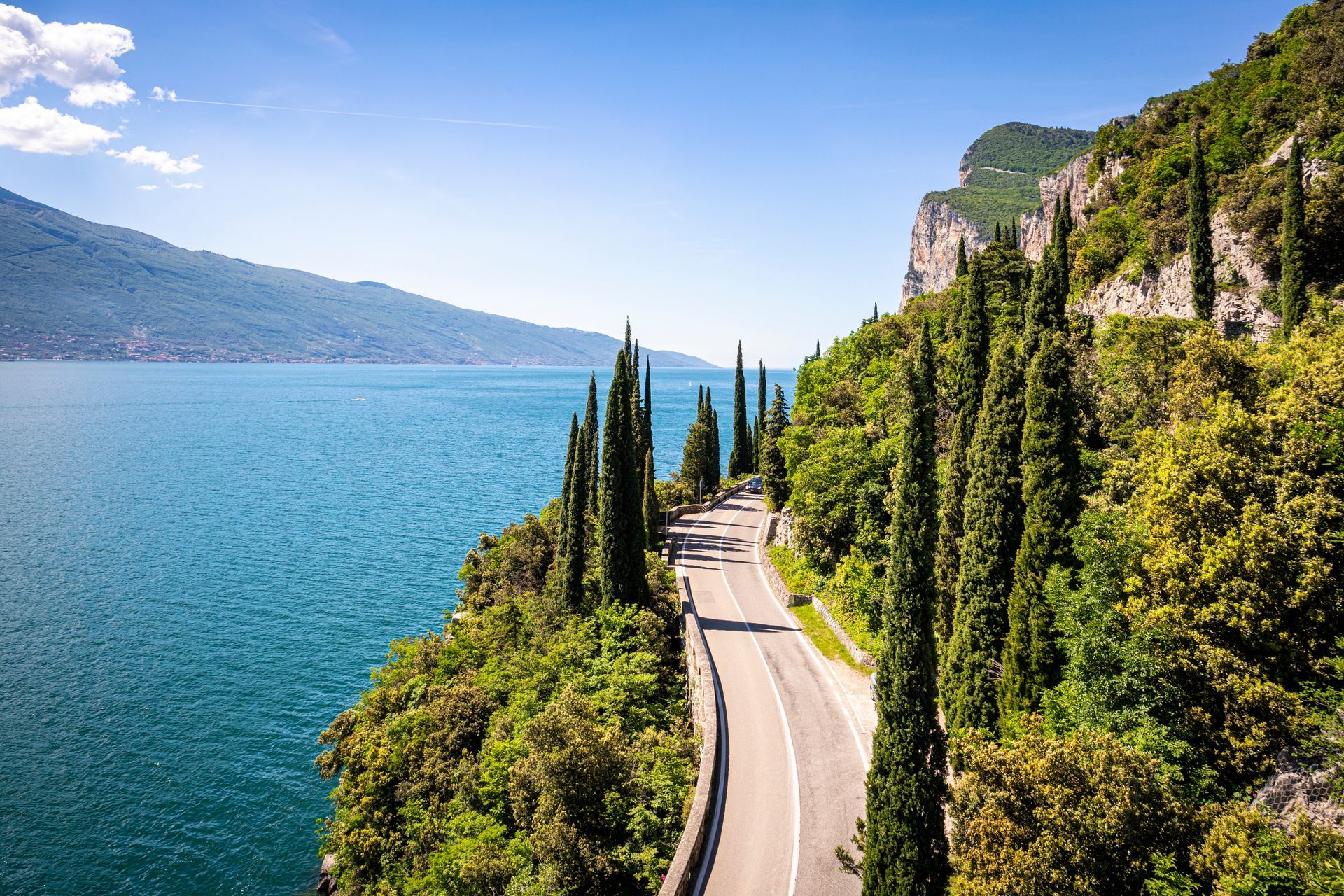 Road winding along a cliffside next to a turquoise lake, flanked by tall trees, under a blue sky.