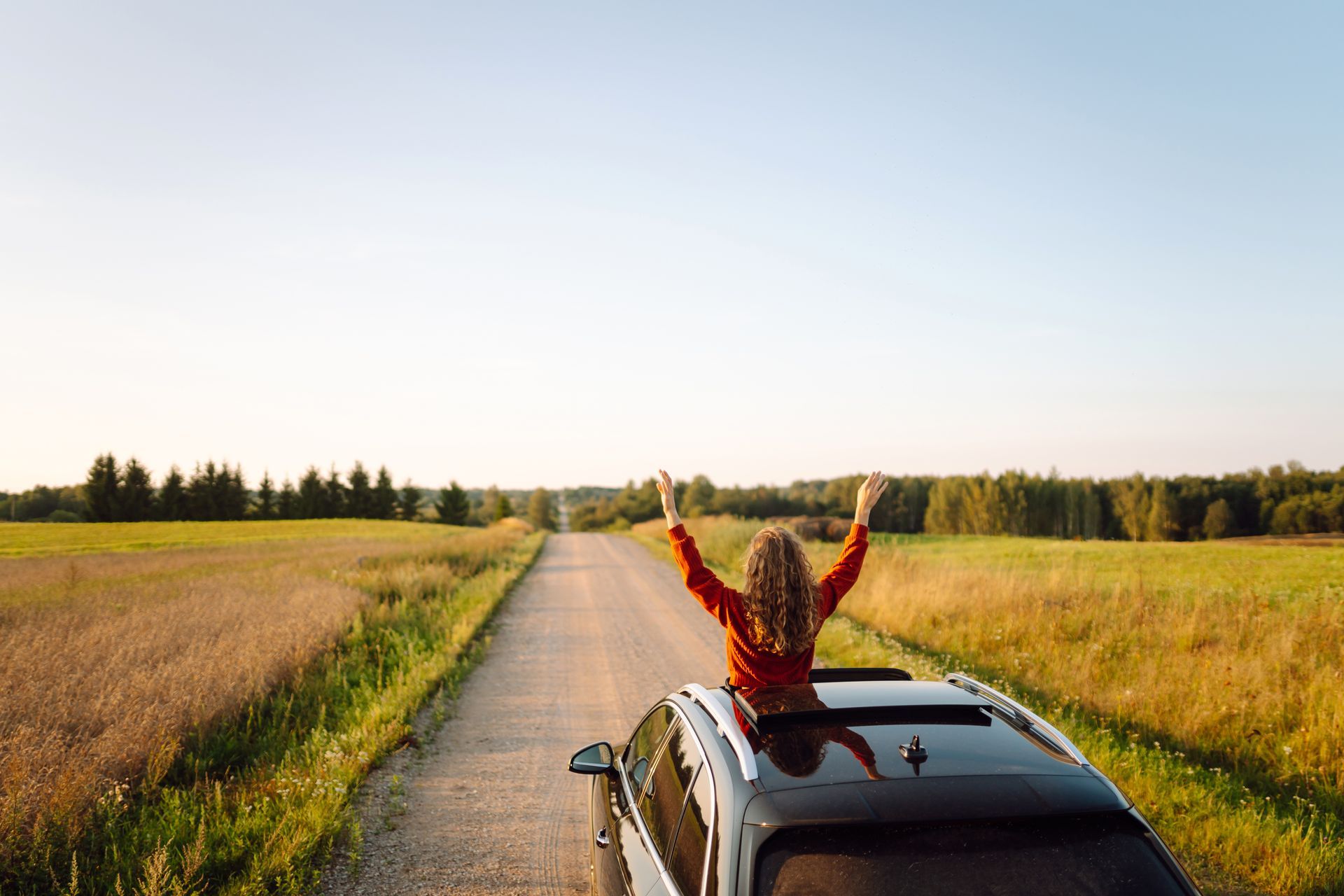 Woman with arms raised through car sunroof on rural road.