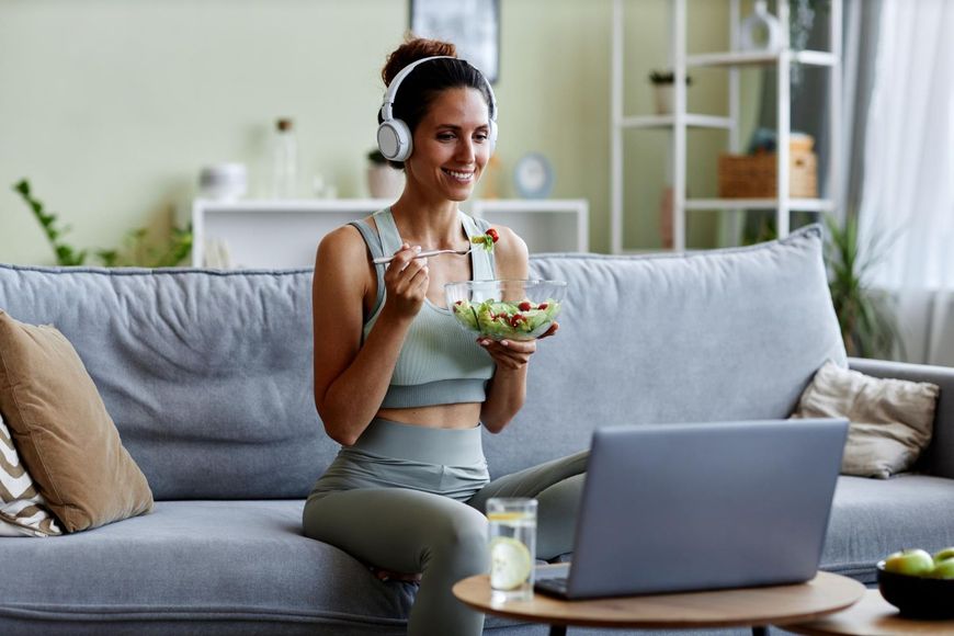 Woman in workout clothes sits on couch, eating salad, using laptop with headphones in living room.