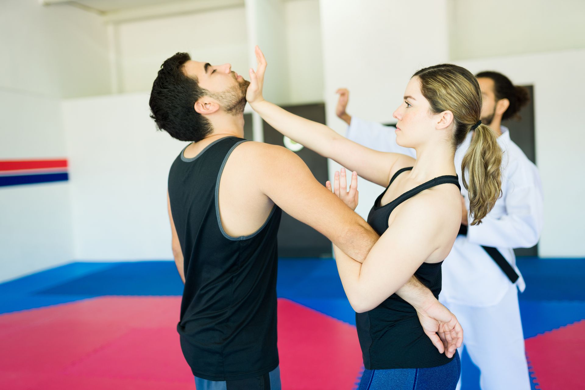 Two people practicing a defensive move in a self-defense class with instructors nearby.