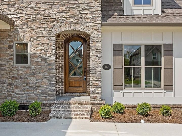 Exterior of a house with stone facade and a wooden arched door. White siding and brown shutters are visible.