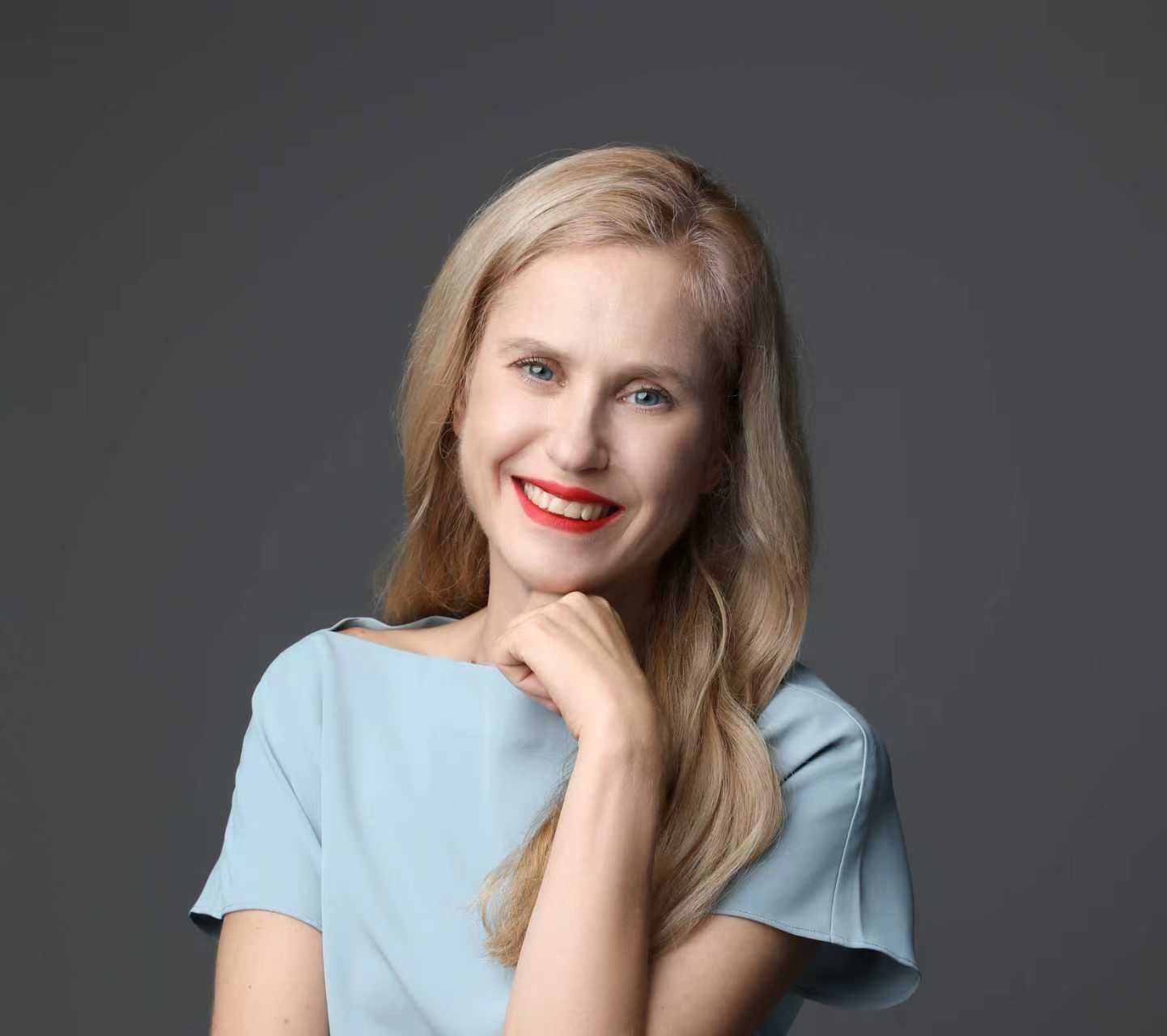 Woman with blonde hair, smiling, wearing a light blue top, resting her chin on her hand, against a gray background.