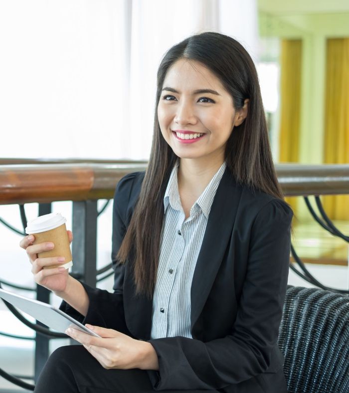 A woman in a suit is sitting on a couch holding a cup of coffee and a tablet.