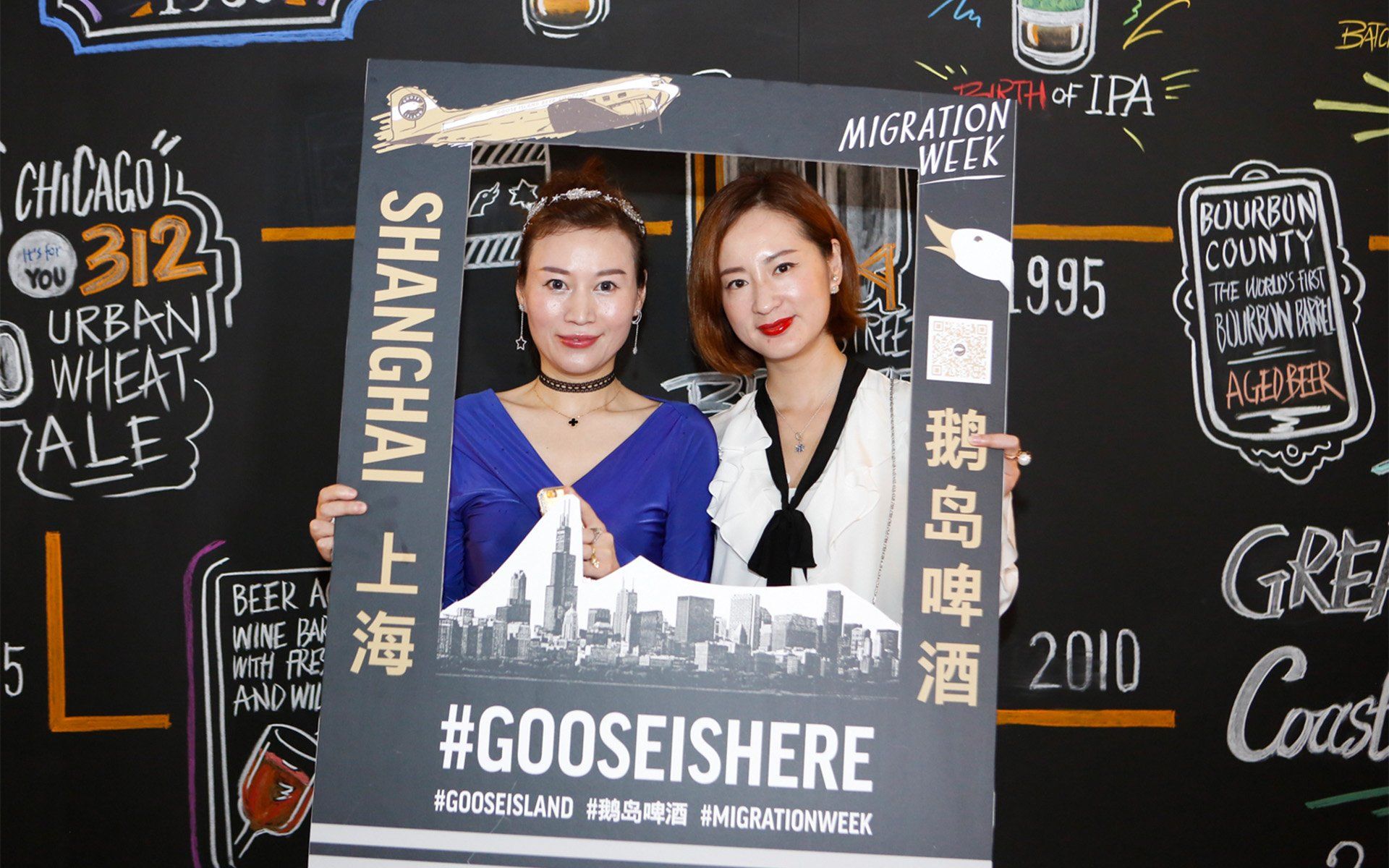 Two women are holding a sign that says gooseishere in front of a chalkboard.