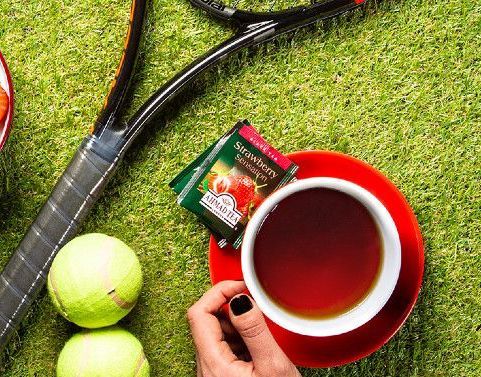 A person is holding a cup of tea next to tennis balls and a tennis racquet.