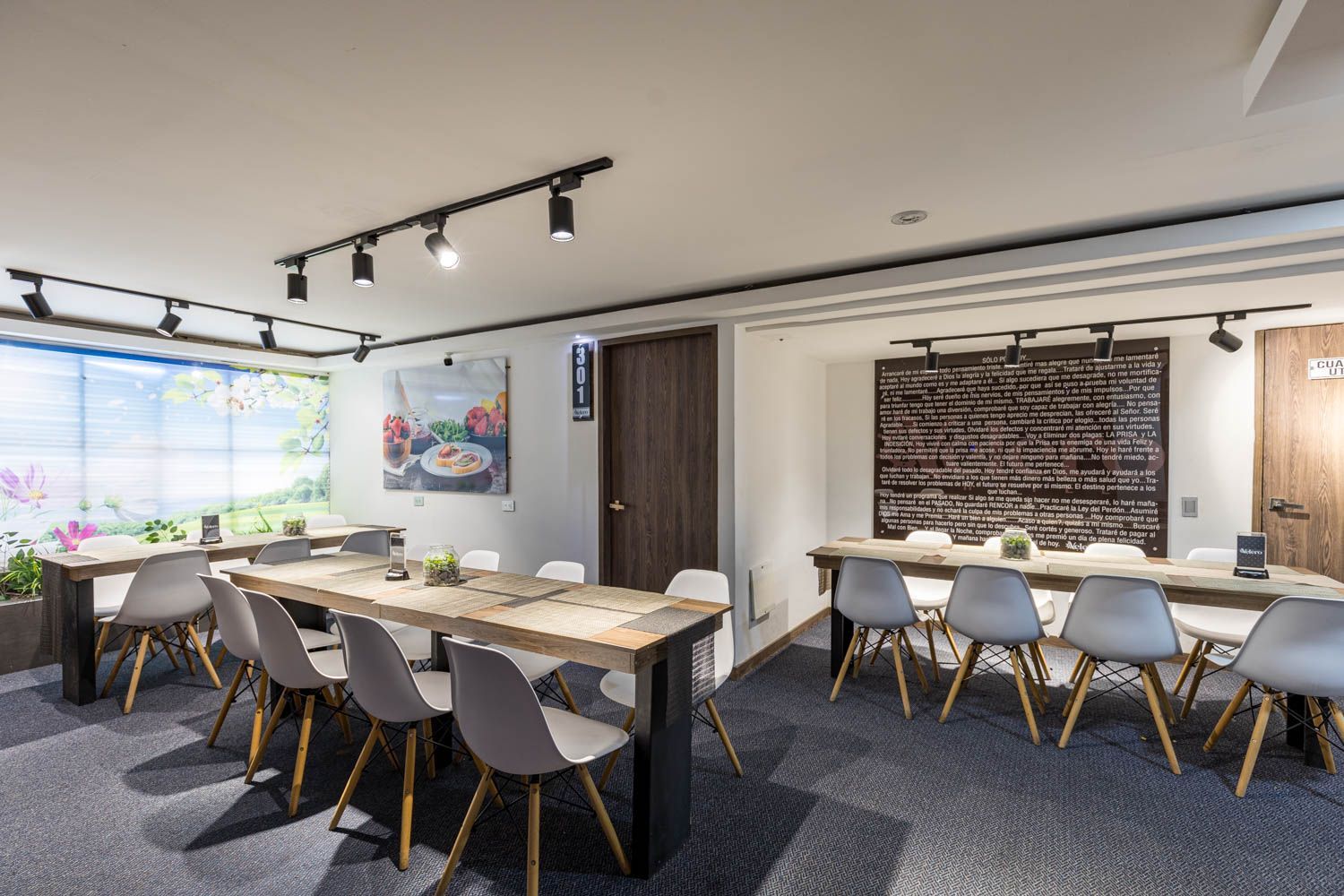 Un comedor moderno con largas mesas de madera, sillas grises e iluminación en riel. Una pared tiene un gran menú y otra una ventana con un mural floral.