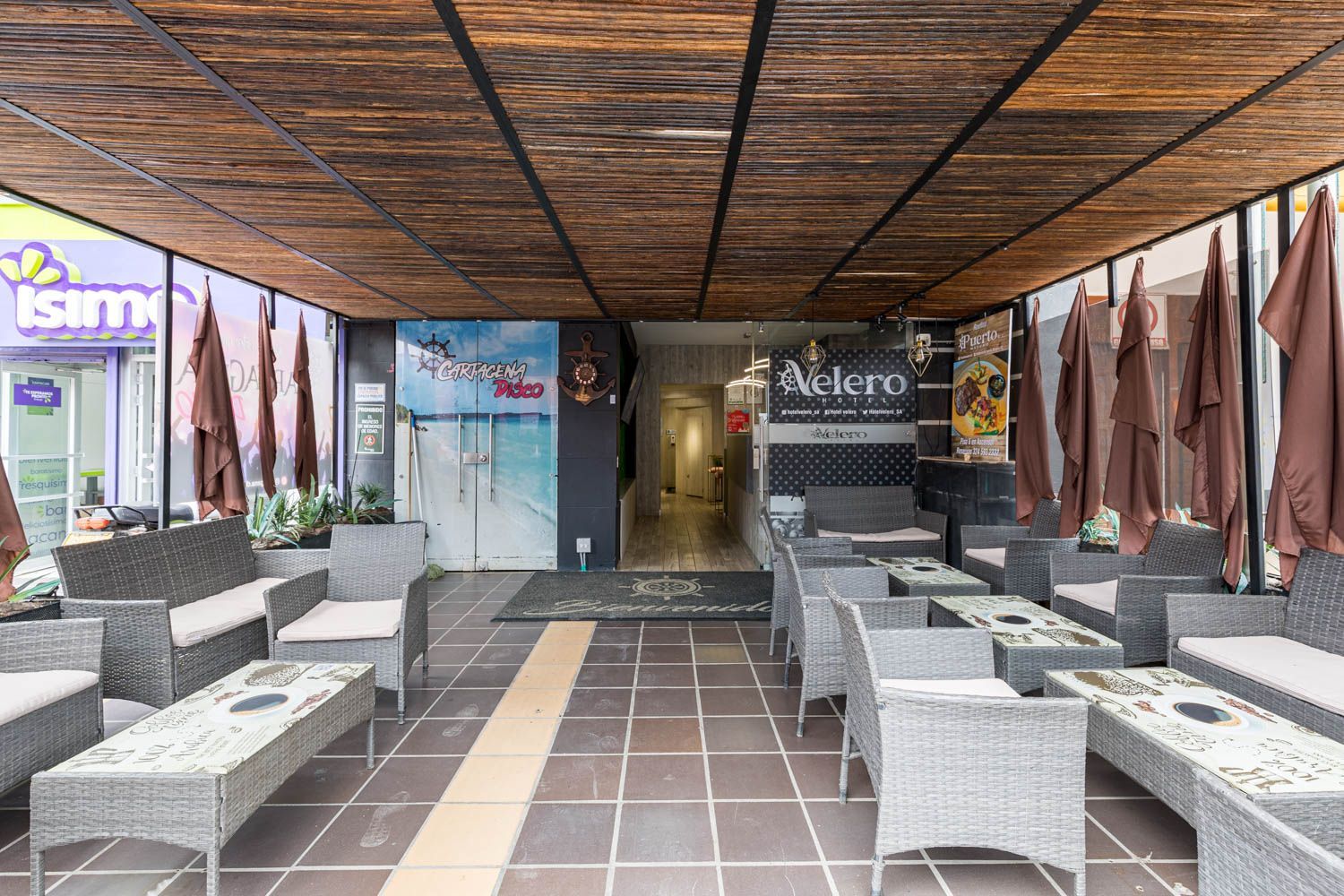 Zona de estar al aire libre de una cafetería, con muebles de mimbre bajo un dosel tejido. Una puerta da acceso al interior.