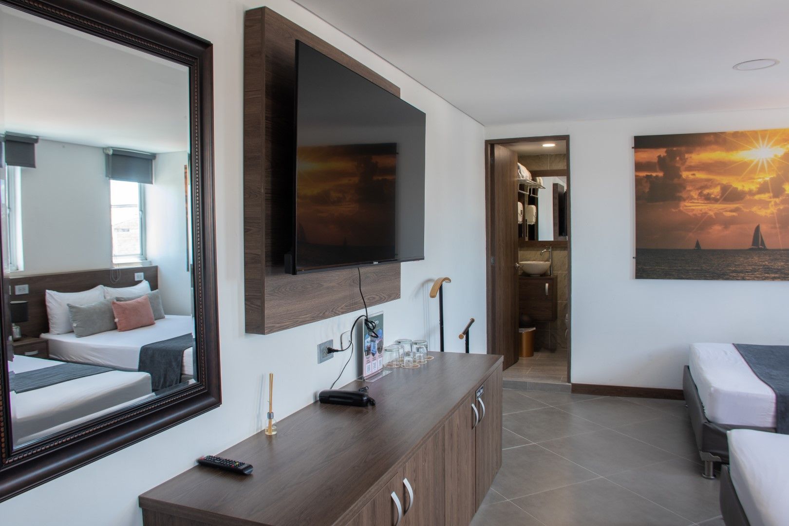 Habitación de hotel con dos camas, espejo grande, TV montada en la pared y puerta abierta a un baño.