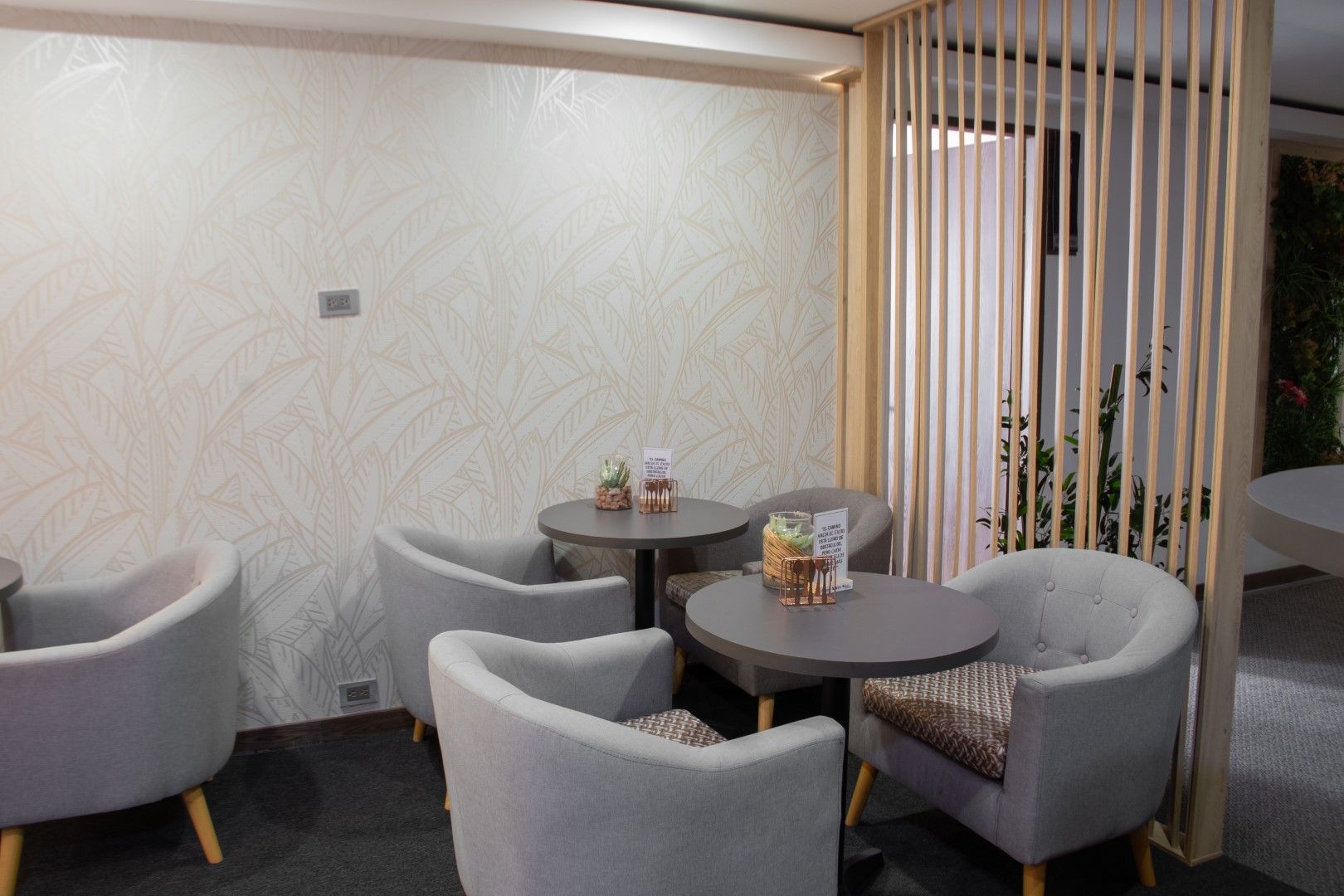 Una moderna zona de cafetería con sillas grises y pequeñas mesas redondas. Un separador decorativo de listones de madera separa parcialmente el espacio.