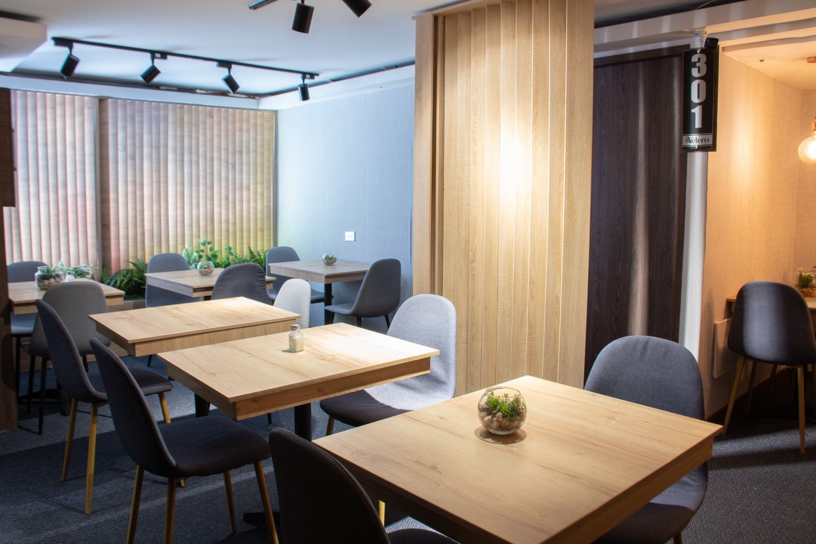 Interior de cafetería moderna con mesas y sillas de madera, colores claros y luz natural. Las mesas tienen plantas pequeñas.