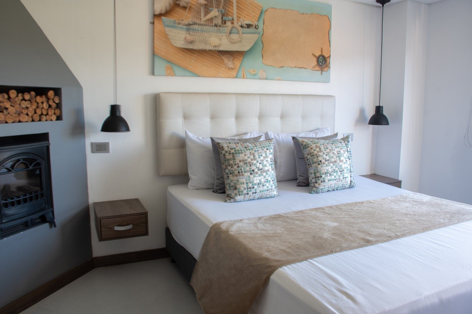 Dormitorio con cama, almohadas, obra de arte de un barco, chimenea y lámparas colgantes. La paleta de colores es neutra, con detalles en madera clara y azul.