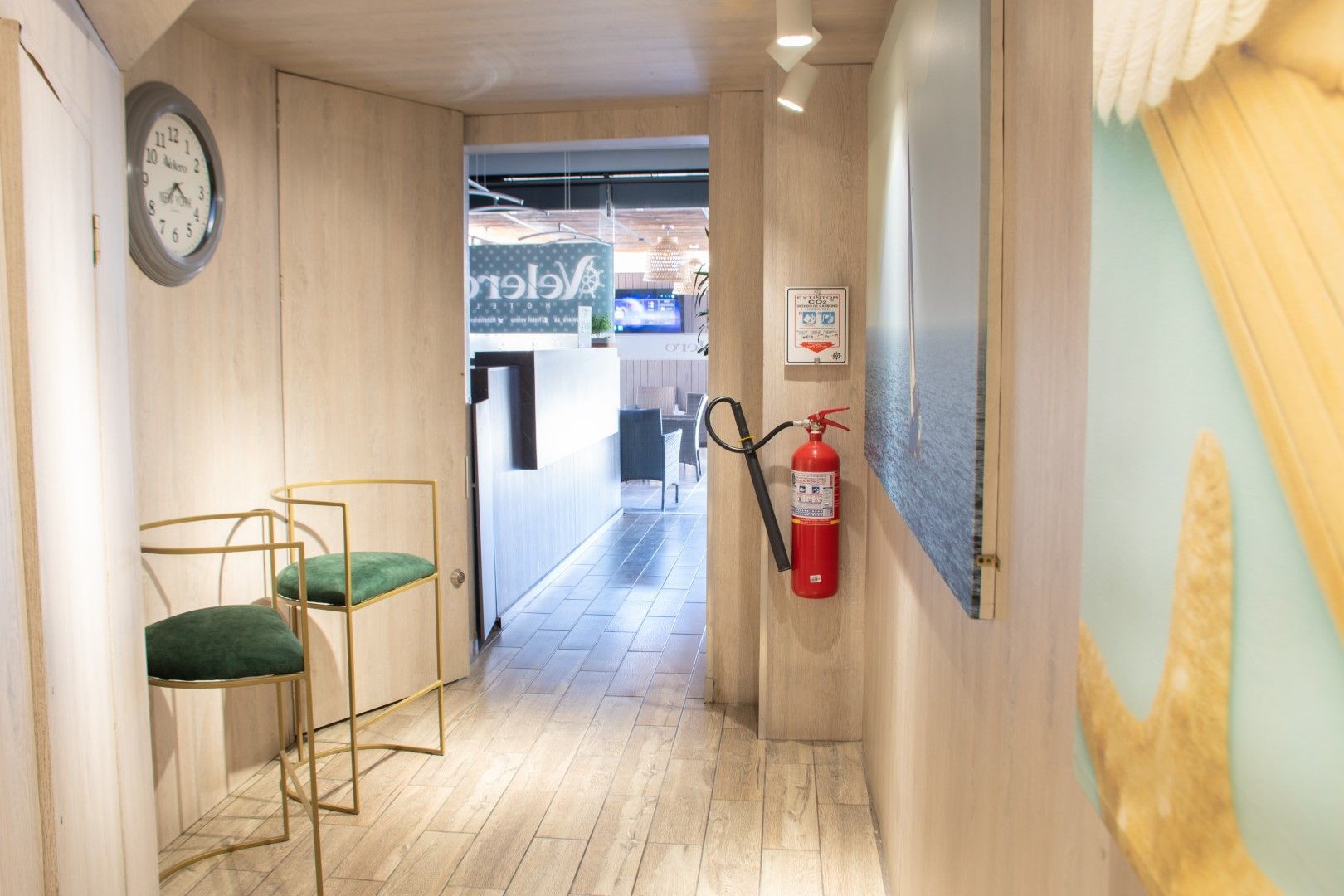 Un pasillo estrecho con paneles de madera clara conduce a una puerta de cristal. Dos sillas doradas con asientos de terciopelo verde se encuentran junto a una pared con un reloj. Un extintor cuelga de la pared.