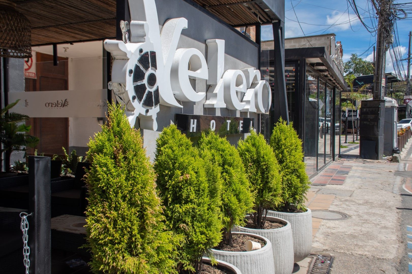 Fachada del Hotel Melezo con un gran cartel blanco y arbustos verdes en la entrada. Edificio con fachada en negro y gris.