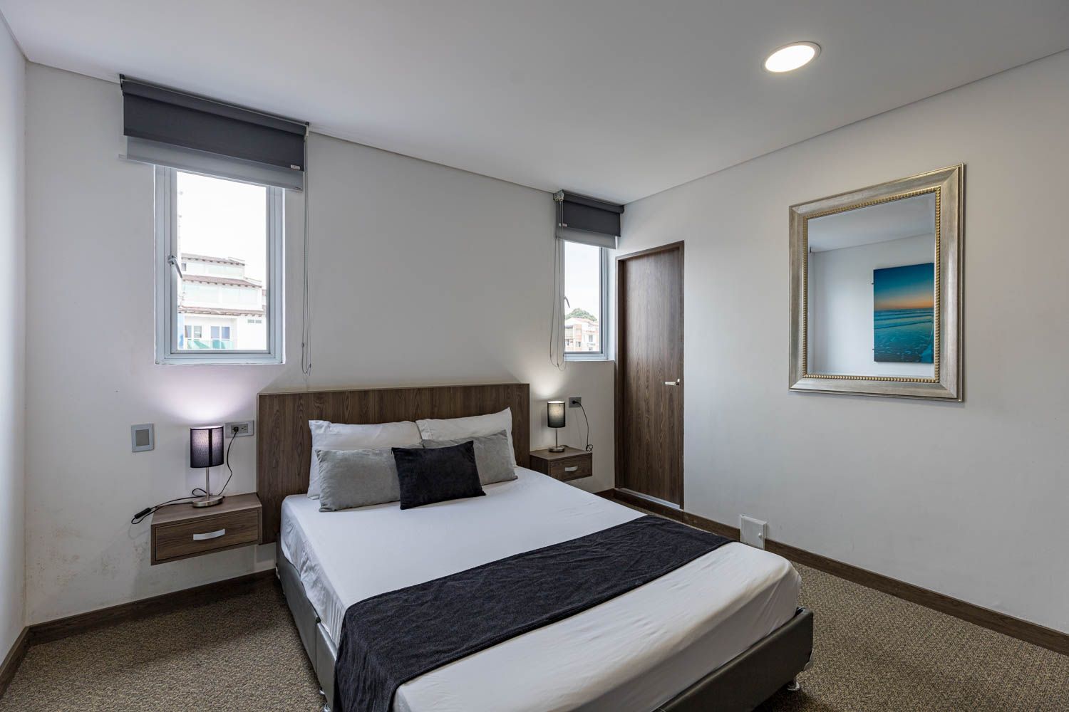 Dormitorio con cama, mesitas de noche, ventanas y espejo. Las paredes son blancas, con alfombra marrón y un cubrecama oscuro.