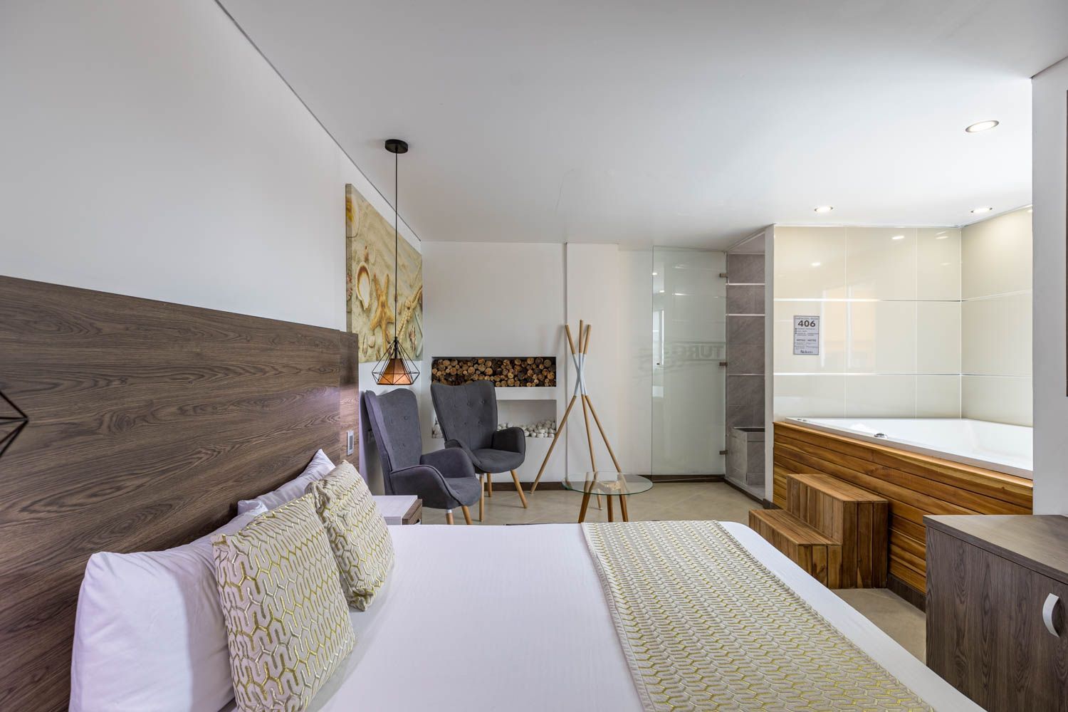 Habitación de hotel con cama, dos sillones, jacuzzi y elementos decorativos. La habitación es luminosa, con colores neutros y detalles en madera.