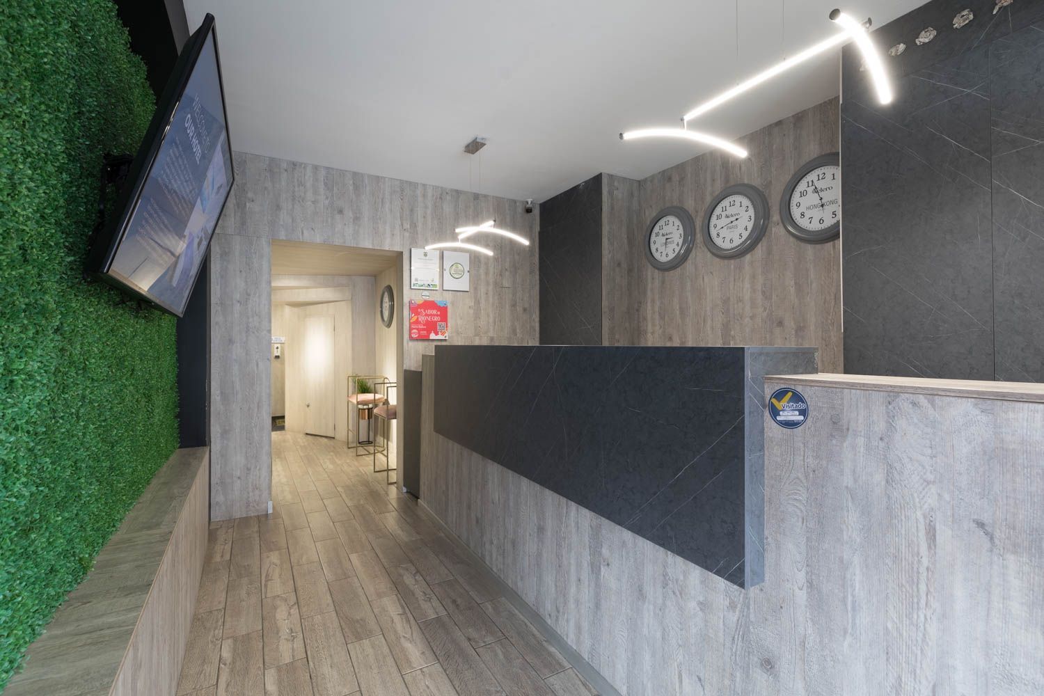 Área de recepción del hotel con paneles de madera gris, un mostrador de recepción negro, una pared verde y relojes mundiales.