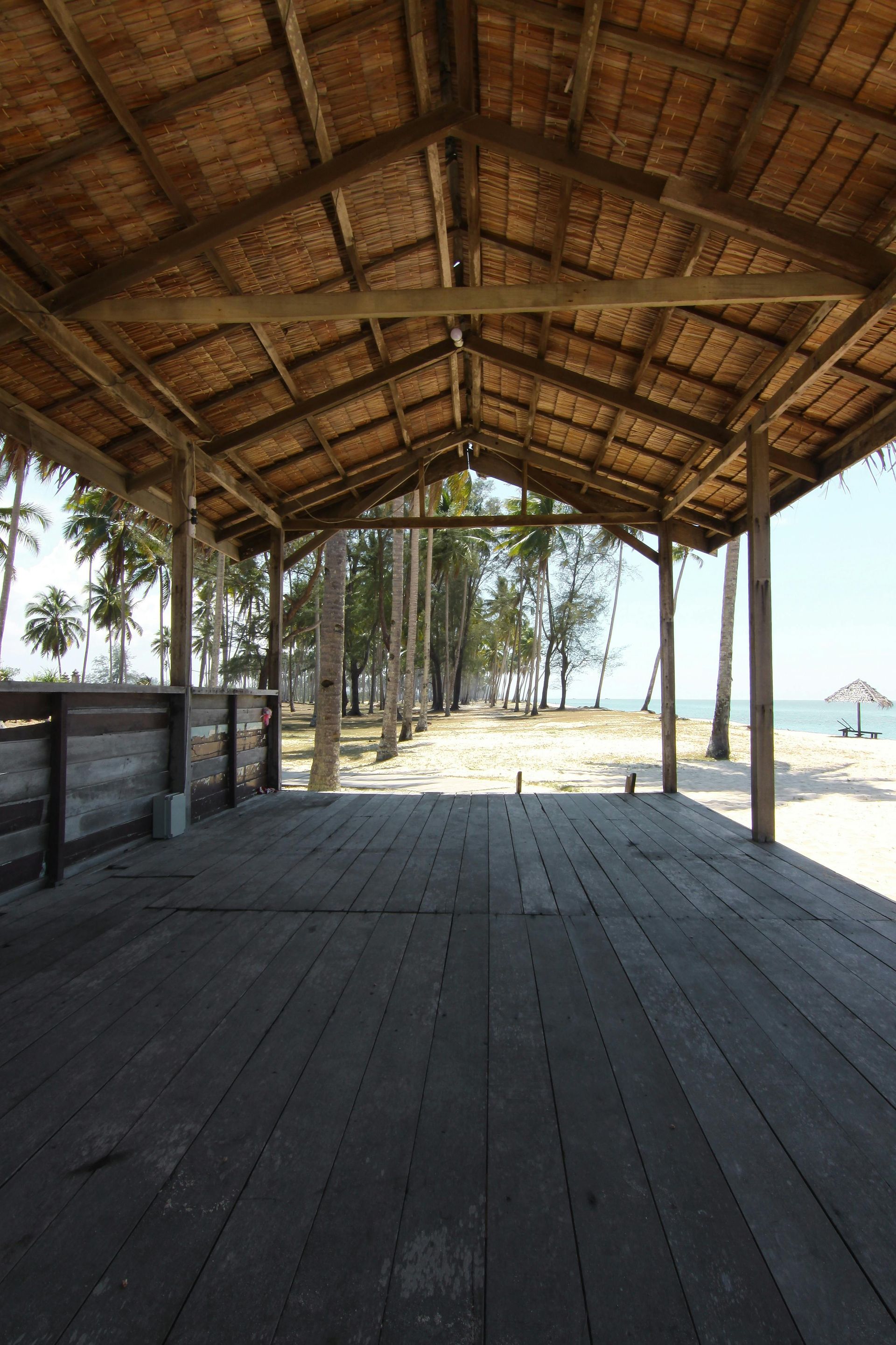 Wooden boardwalk under a thatched roof structure, leading to a beach with trees and a view of the ocean.