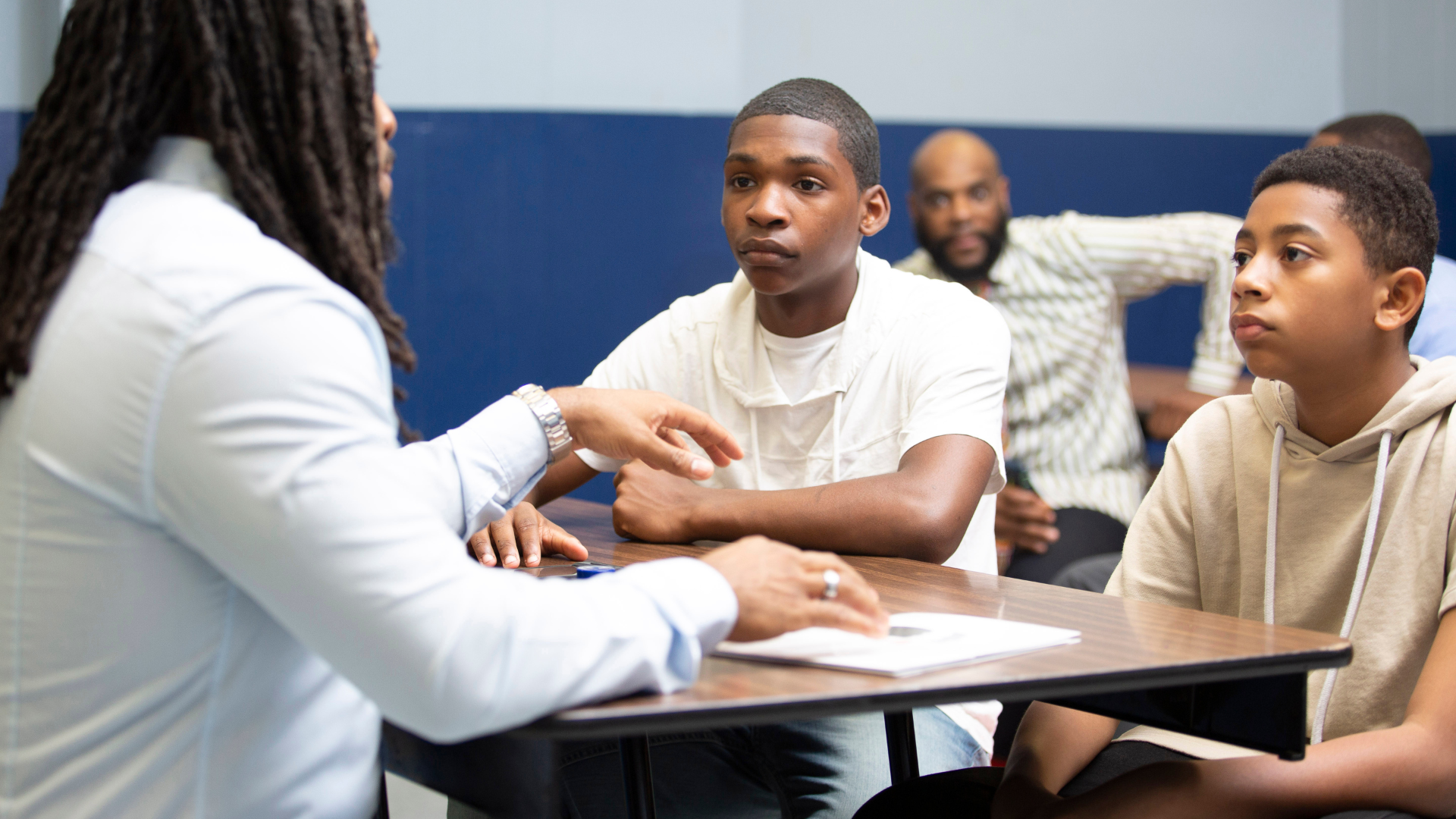 A group of young men are sitting at a table talking to a woman.