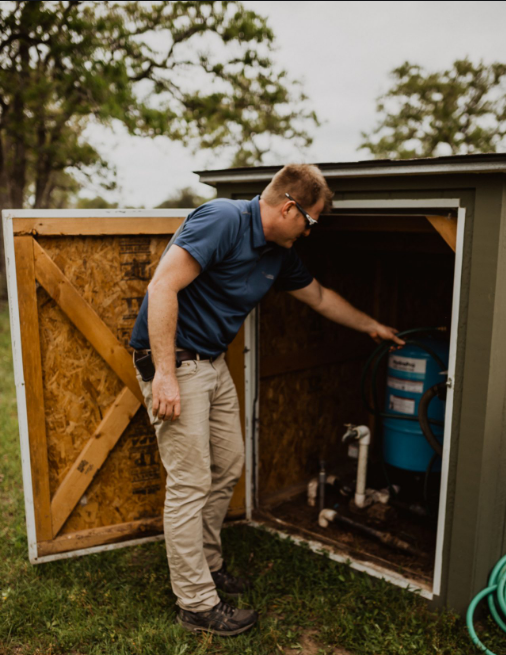 Man inspecting water pump inside a small shed outdoors; he's wearing glasses, a blue shirt, and tan pants.