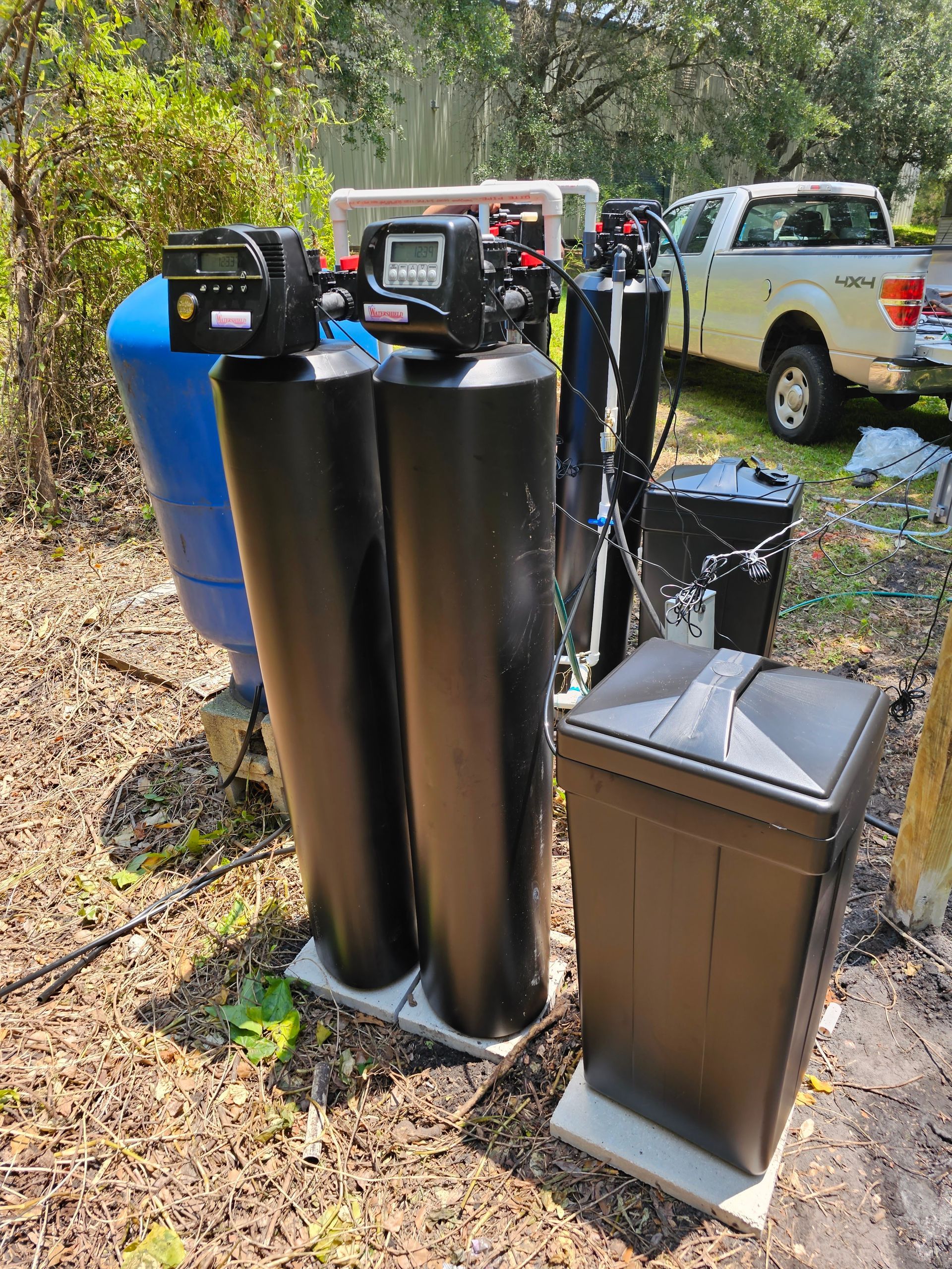 Water filtration system with black and blue tanks, digital control panel, and a trash can, outdoors near a truck.