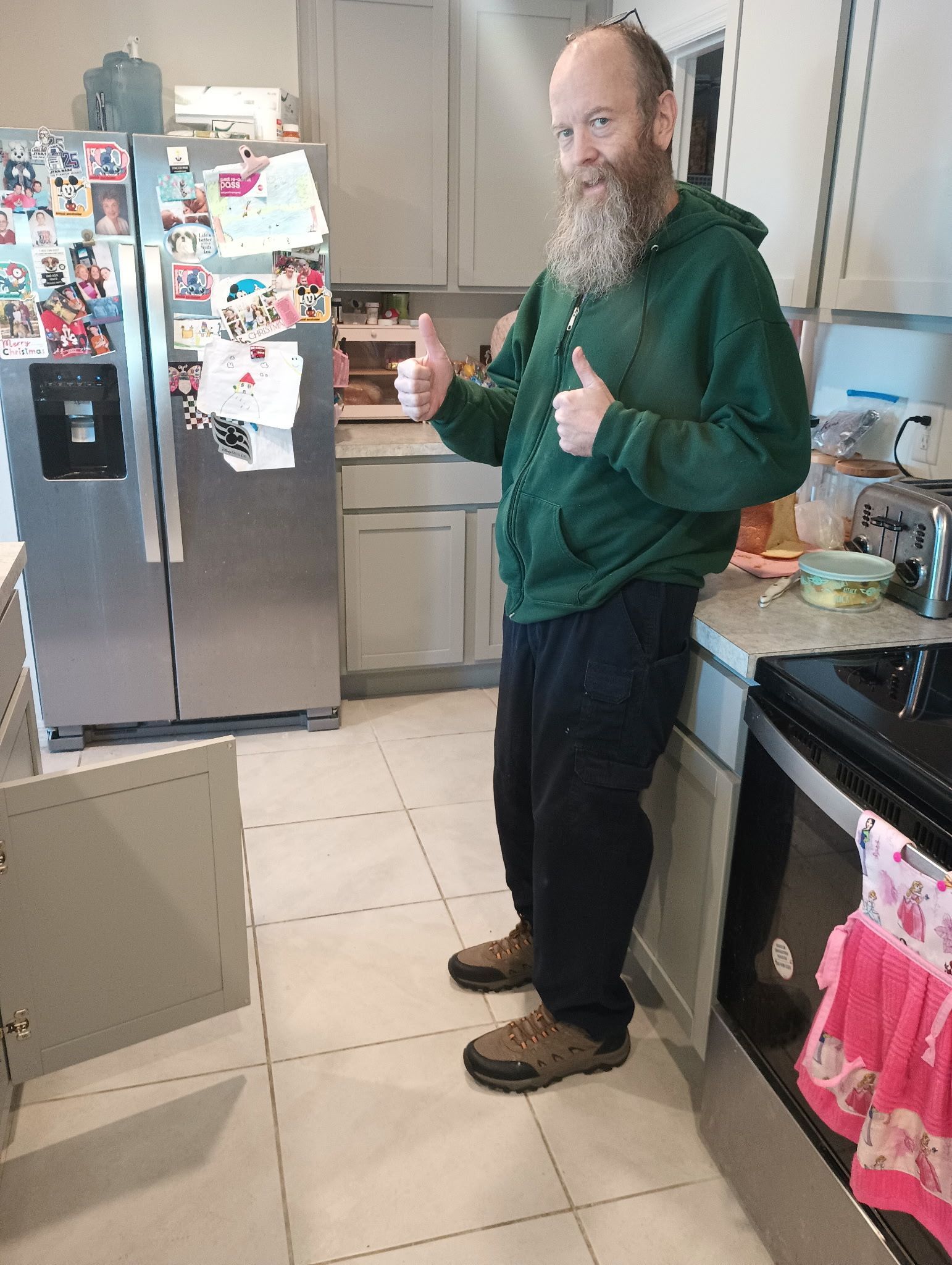 Man in green hoodie, giving thumbs up in a kitchen with open cabinets and a refrigerator.