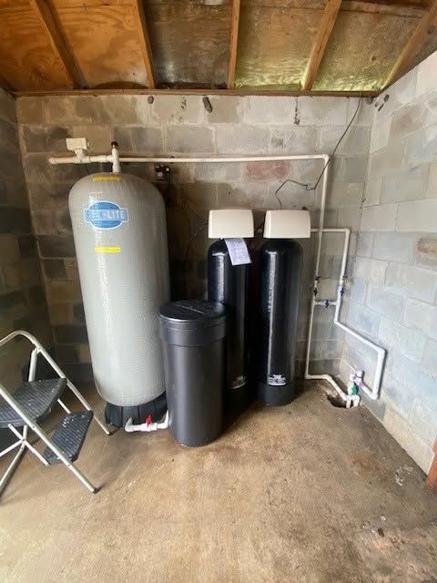 Utility room with water softener tank, black filter canisters, folding chair, and cinder block walls