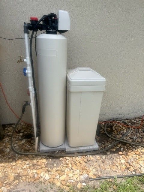 A water softener system, comprising a tall white tank, a smaller white salt tank, and piping, is set outside on gravel.