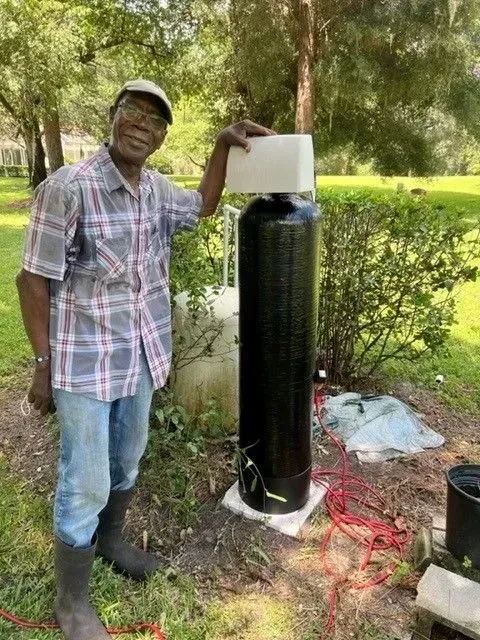 Man standing beside a black cylindrical tank outdoors, holding the top in a grassy area with tools nearby