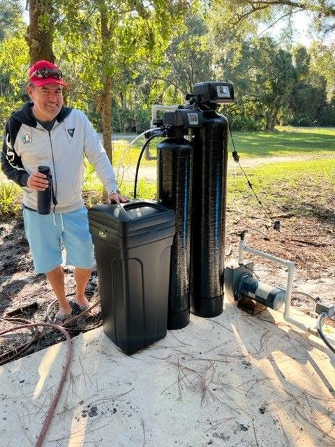 Man standing next to a water filtration system outdoors; trees in the background.