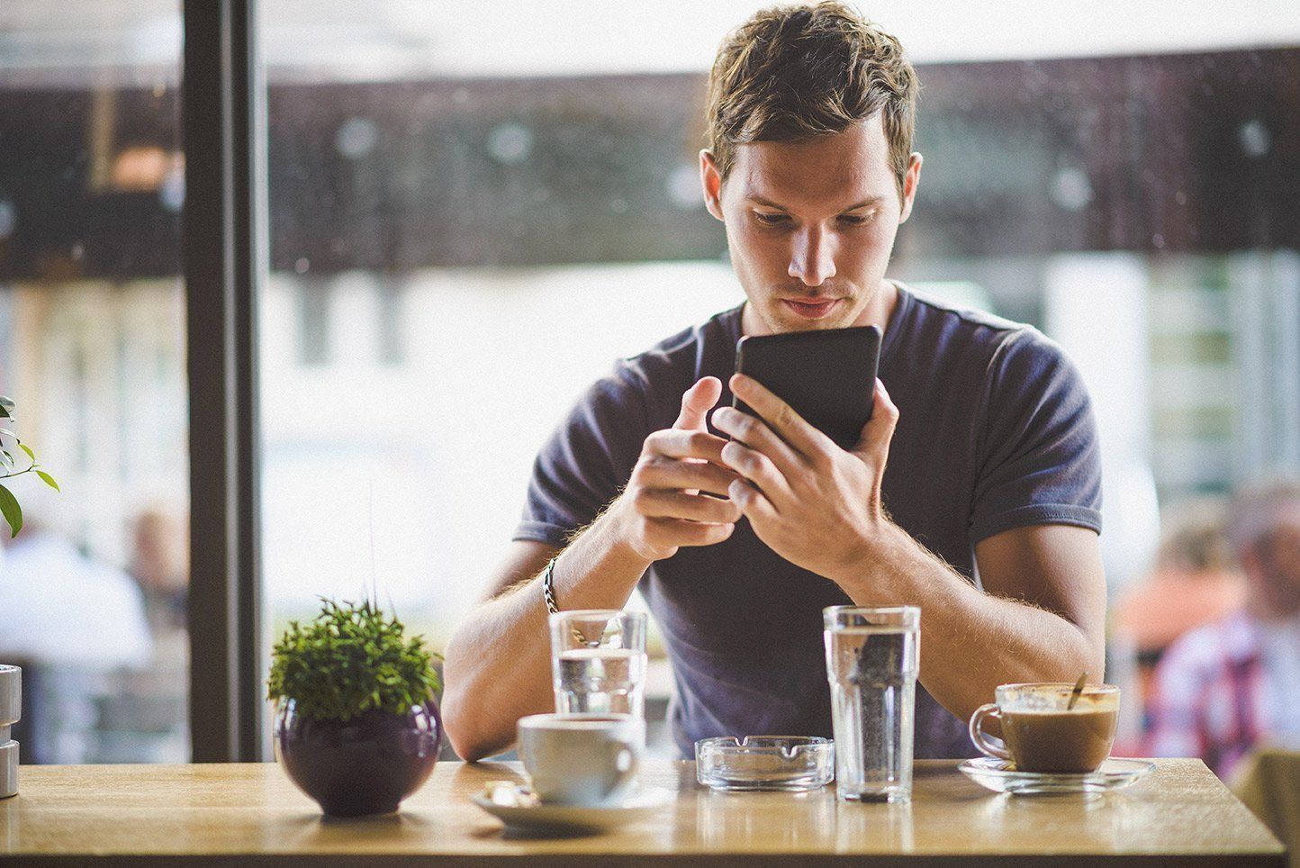 Man looking at a phone while sitting at a table in a cafe, with drinks and a small plant.