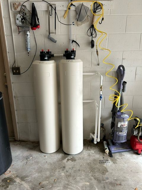 Two white water filtration tanks in a garage setting with equipment hanging on the wall and a vacuum cleaner.