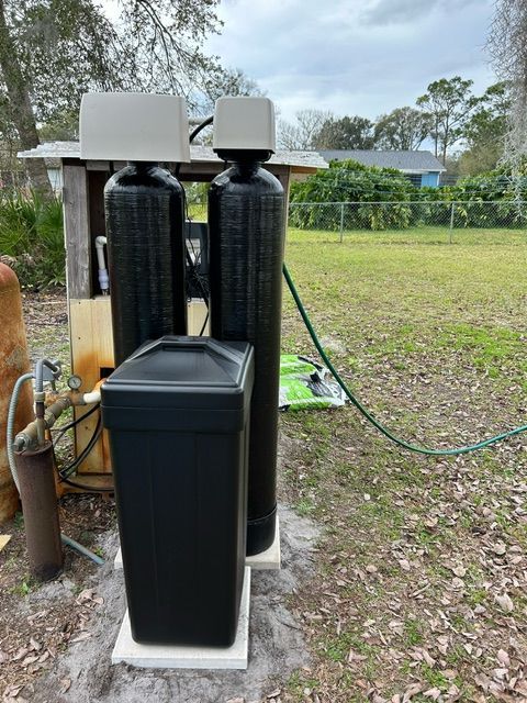 Water softener system with two black tanks and a salt storage tank outdoors. Green hose and grass visible.