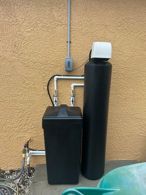 Water softener system installed on an exterior stucco wall. Black tanks with white plumbing.