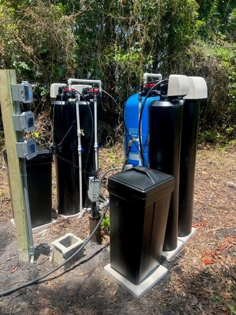 Water filtration system with black tanks, blue pressure tank, and black control boxes on concrete pads outdoors.