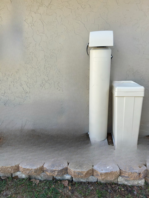 Water softener system on a concrete pad, beige tank and tall cylinder against stucco wall.