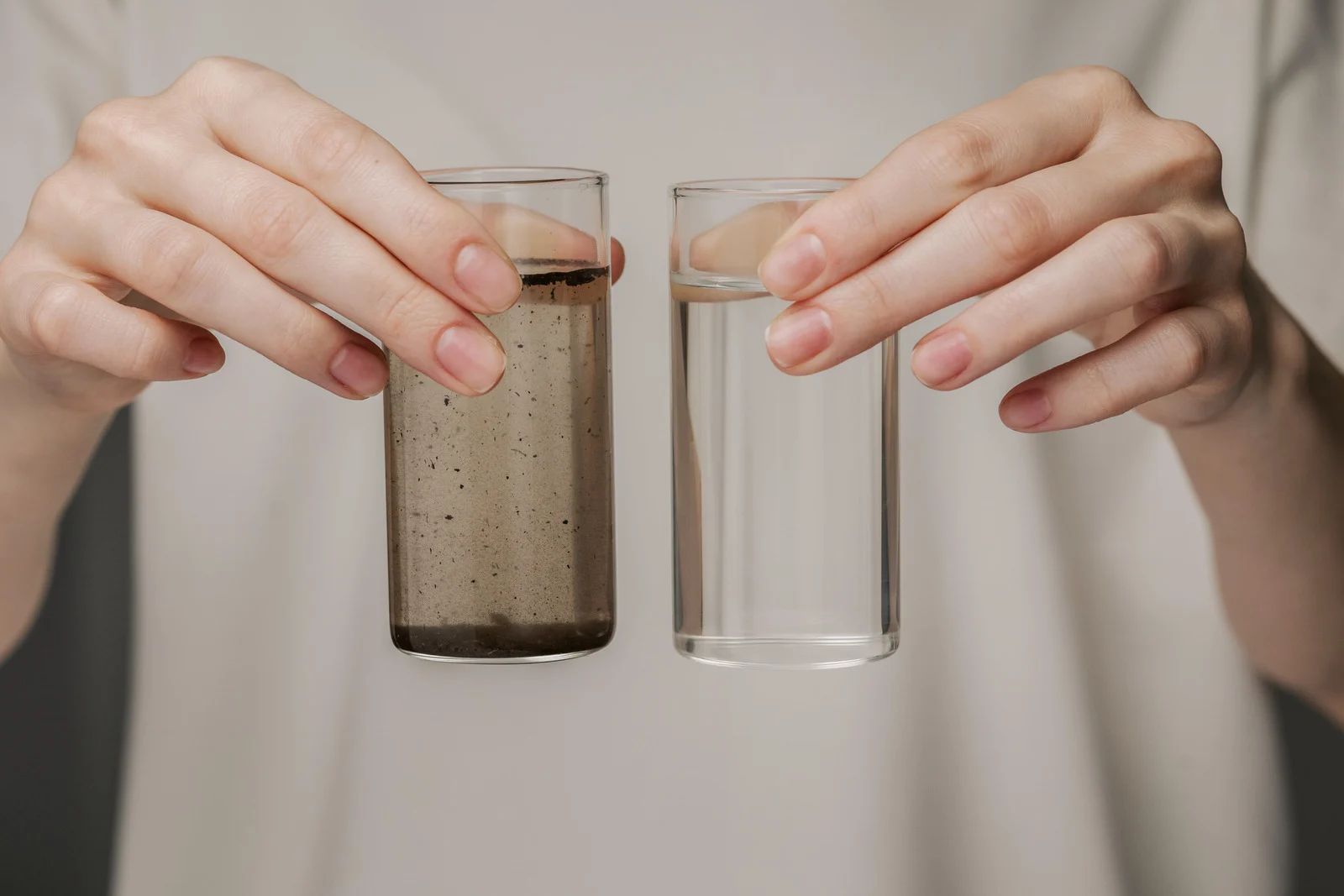 A person holds two glasses of water. One glass is clear, the other is murky brown