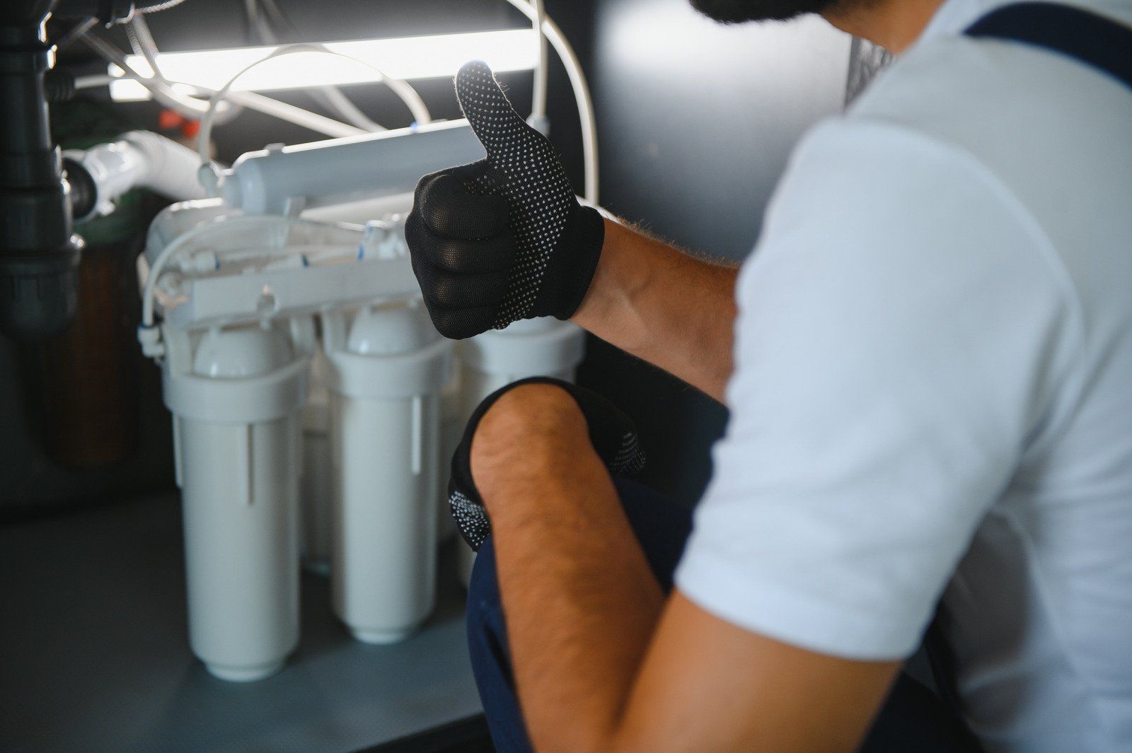 Plumber installing a water filter under a sink; gives thumbs-up, wearing gloves.