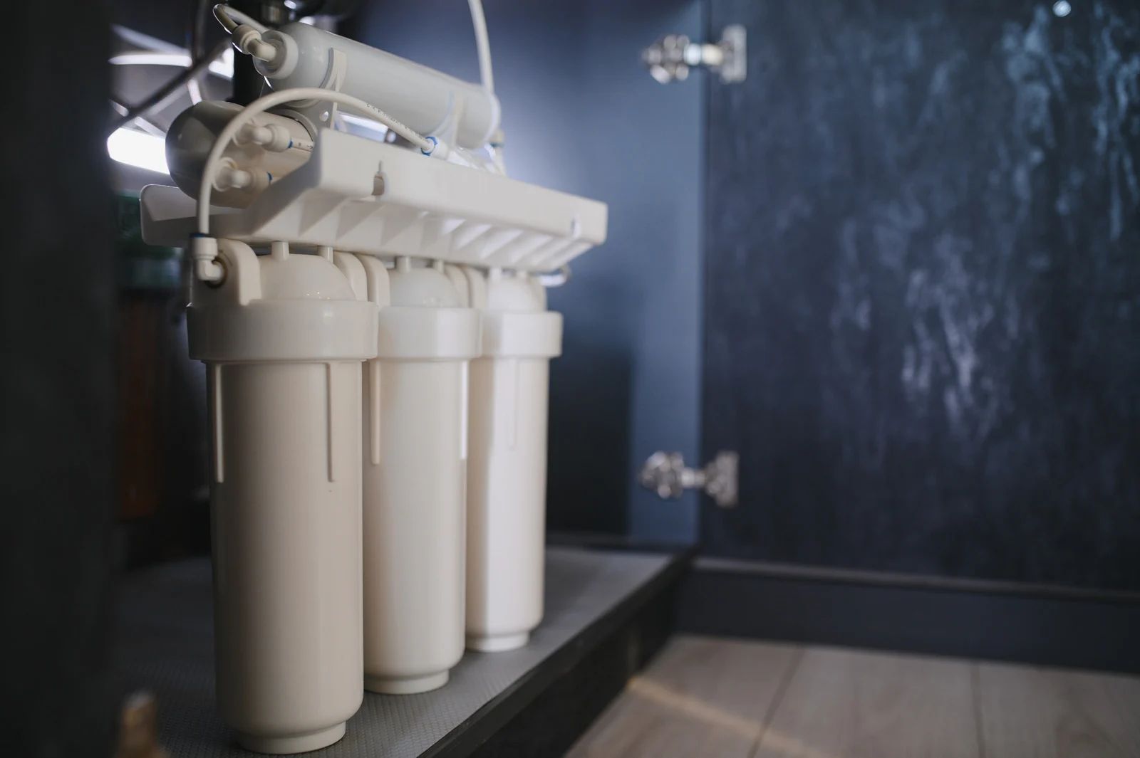 Water filter system inside a dark cabinet with three white cylindrical filters.