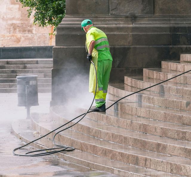 Worker in neon green gear pressure-washing stone steps, with mist and hose on a city sidewalk