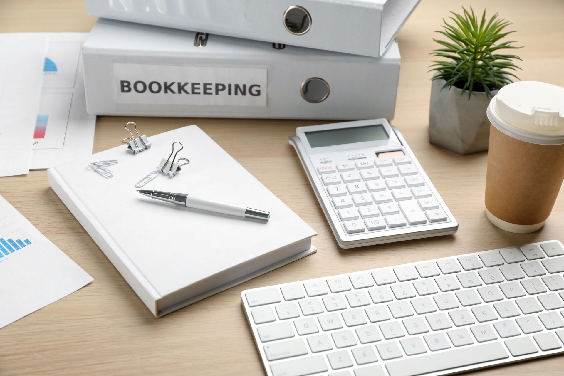 A desk with binders , a calculator , a keyboard , a pen , and a cup of coffee.