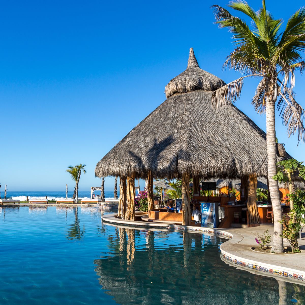 Poolside bar with thatched roof next to blue pool, palm trees, and ocean under a sunny sky.