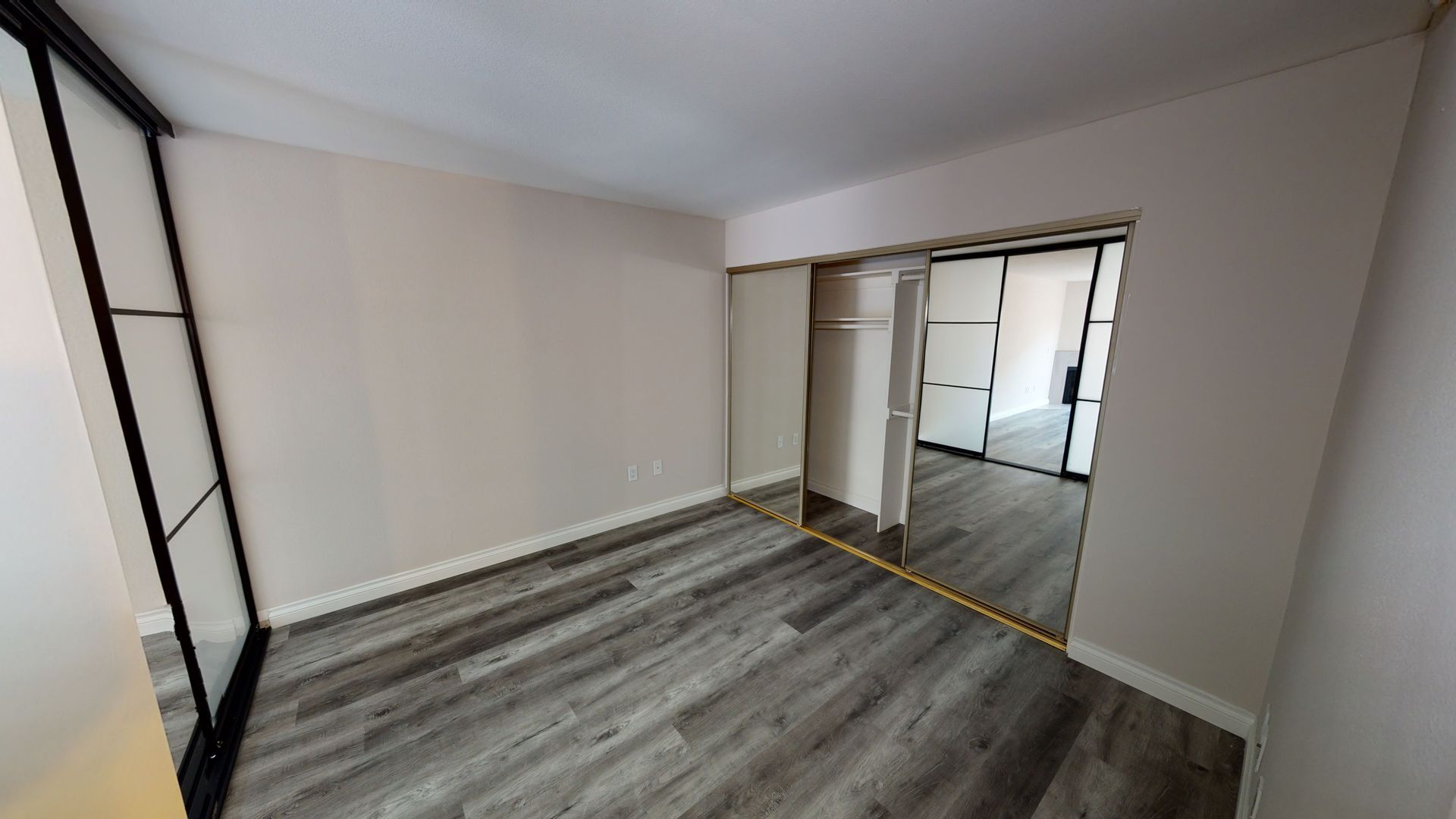 Empty room with gray wood-look flooring, beige walls, sliding mirrored closet doors, and a black-framed window.