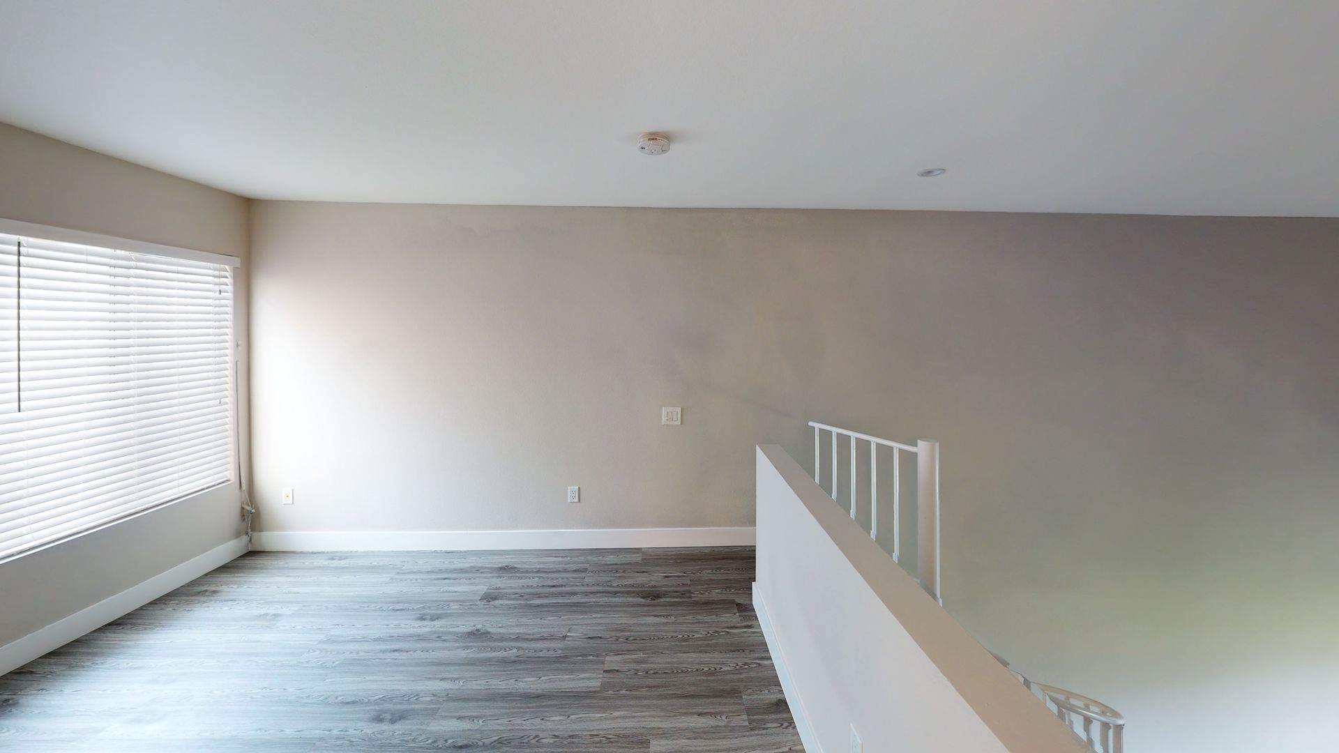 Interior of a loft with a gray floor, white railing, and a window with blinds.