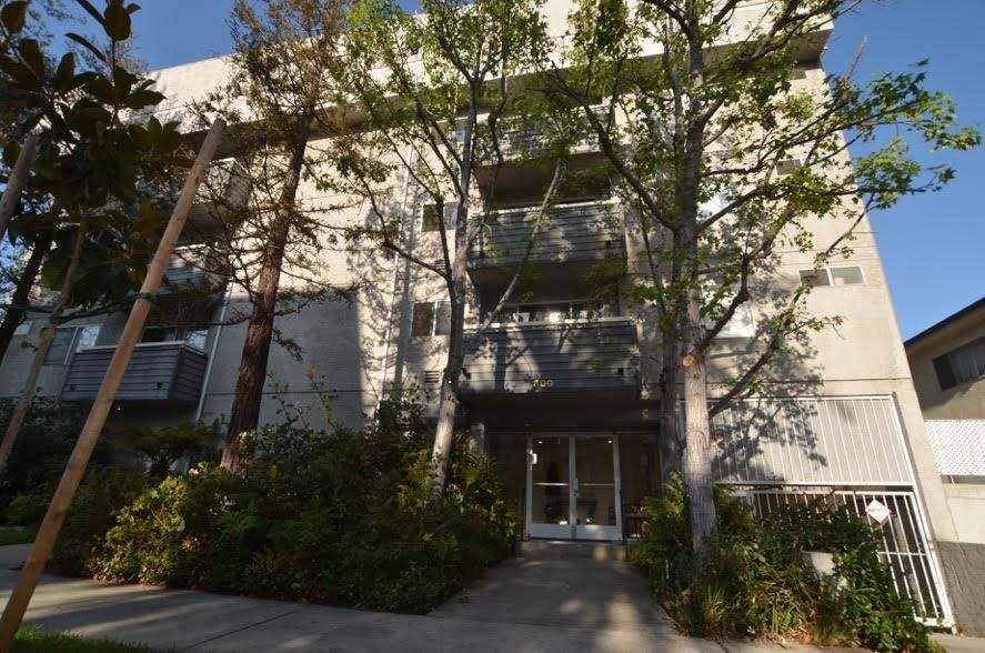 Gray apartment building with balconies, trees, and a walkway.